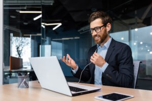 Video call successful businessman boss beard in business suit on online meeting, talking with colleagues using headset and laptop, for remote conversation, man working inside modern office building.