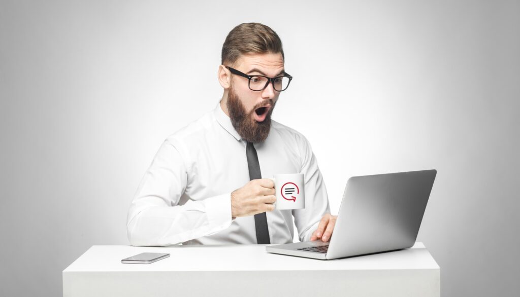 A man is drinking coffee out of a mug featuring the Readback logo. He is using a laptop and looks shocked.