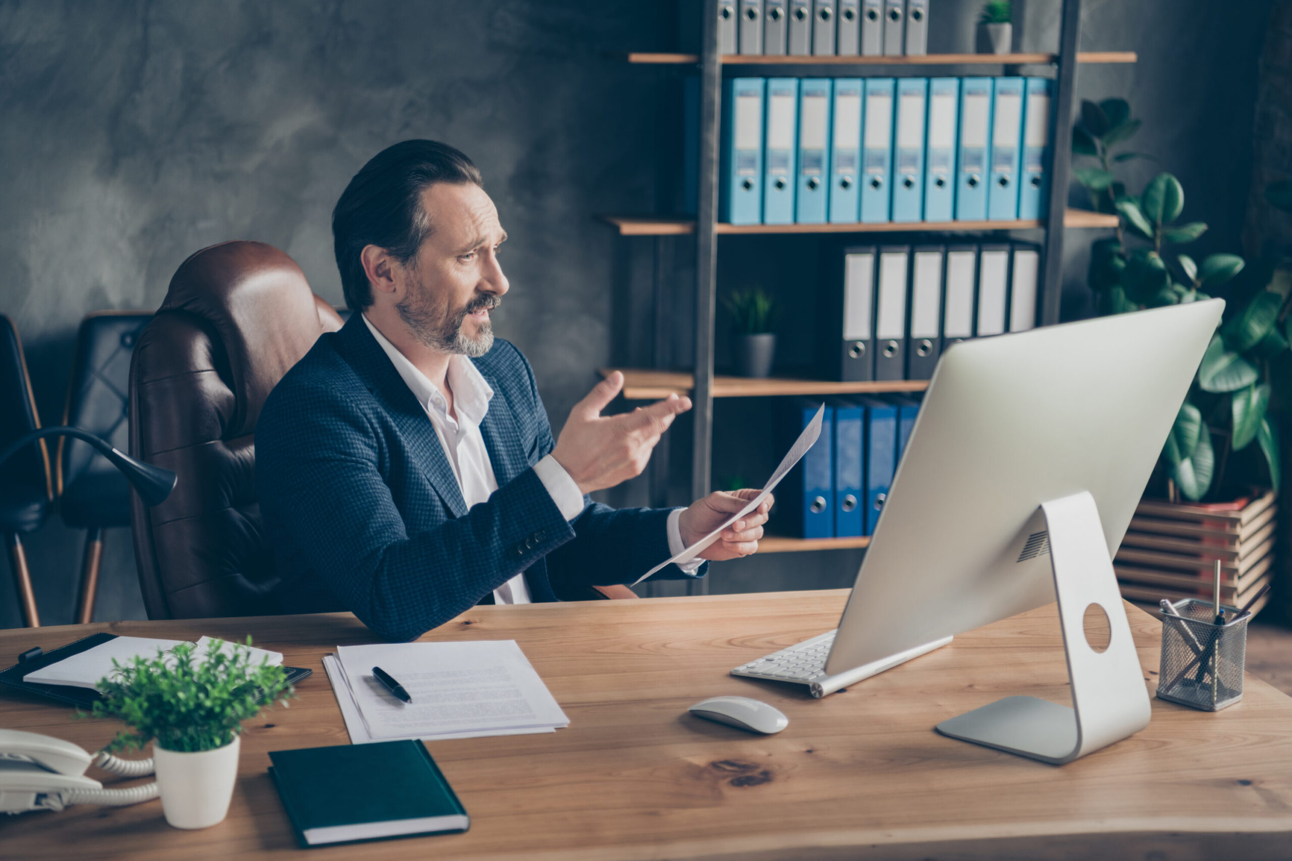 The pictures depicts a man in an office sitting at a desk. The man is in a suit and appears to be in a video meeting. He is holding a piece of paper and gesturing.